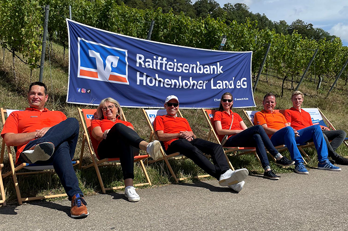 Fünf Mitarbeitende der Raiffeisenbank Hohenloher Land eG machen es sich vor dem Banner entspannt in Liegestühlen bequem und genießen gemeinsam die Genießerwoche.